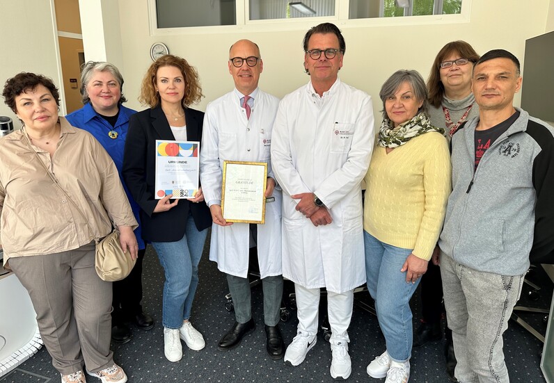 Eine gruppe Menschen steht in einem Raum, zwei von ihnen tragen Arztkleidung. Ein Mann und eine Frau in der linken Bildmitte halten zwei Urkunden in die Kamera.