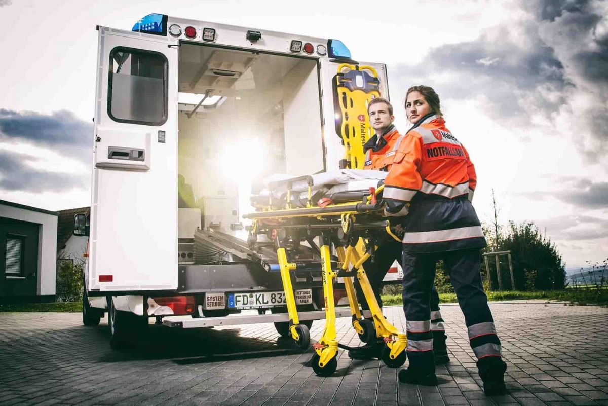 Dramatisch belichtete Szene in der zwei Rettungskräfte eine leere Liege in einen Krankenwagen einladen.