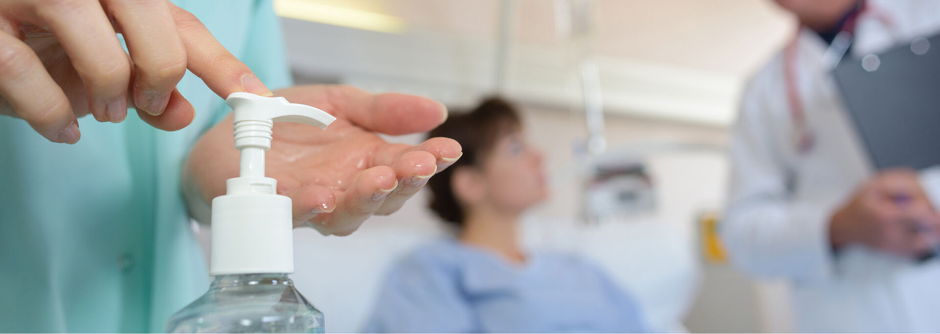 Eine medizinische Fachkraft desinfiziert sich die Hände, während eine Patientin im Hintergrund mit einem Arzt spricht.